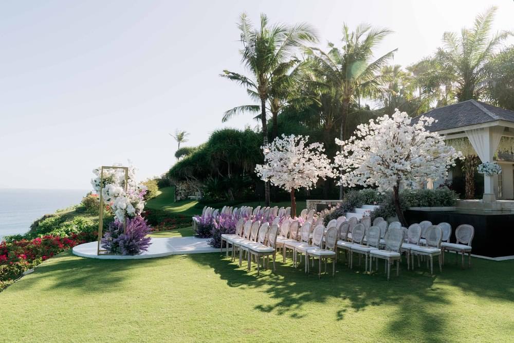 Wedding ceremony setup at Ungasan Clifftop Resort, featuring elegant upholstered chairs, cherry blossom trees, and a floral frame with panoramic ocean views.