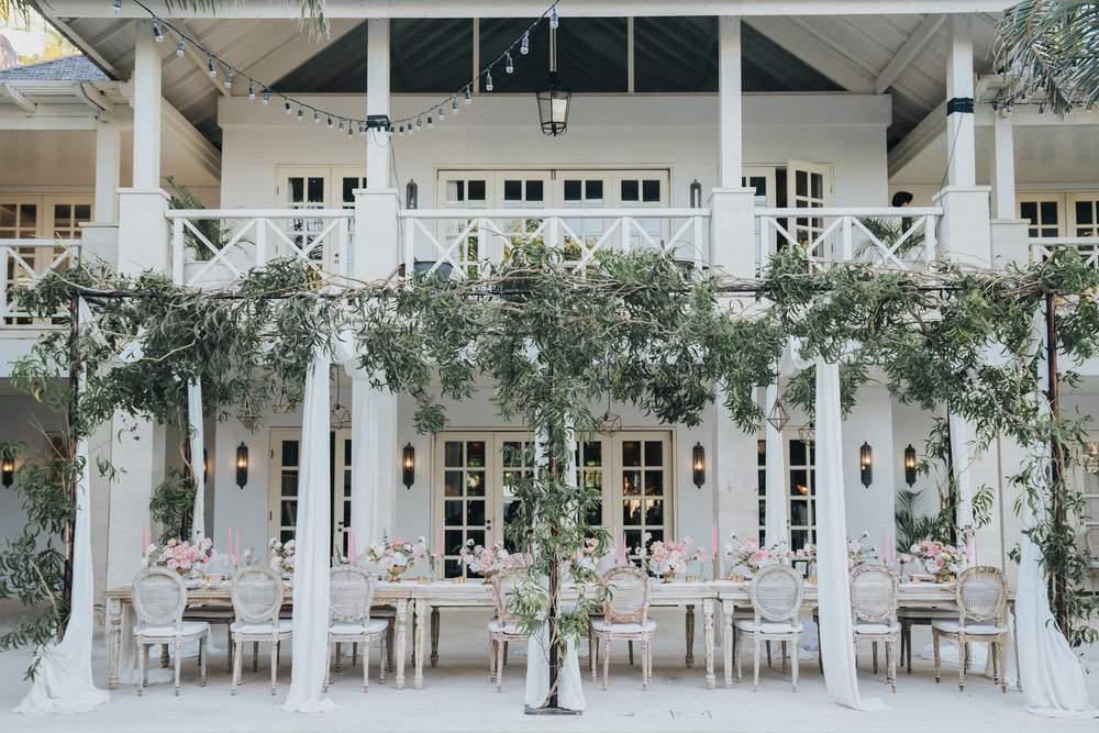 legant wedding reception setup at Ungasan Clifftop Resort, featuring a long dining table with upholstered chairs, draped greenery, and floral arrangements in front of a colonial-style villa.
