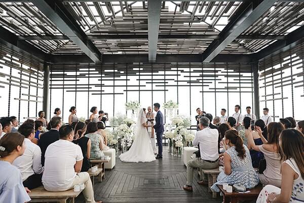 Elegant wedding ceremony setup inside the cabana with white floral arrangements at Alila Villas Uluwatu, Bride and groom being watched by seated wedding guests .