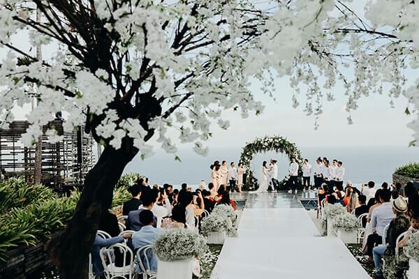 Elegant wedding with bride and groom and seated guests either side of the aisle framed by blooming white floral arches, leading to a floating stage with an ocean-view backdrop at Alila Villas Uluwatu.